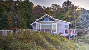 A house with a large bay window, a deck, and a view of the surrounding forest. - Lakefront Montague Cottage w/ Deck & Beach! (Montague)