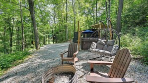 A forested area with a gravel path, a fire pit, Adirondack chairs, and a swing chair. - Tiny Home w/ Hot Tub & Mini Highland Cows! (Loudonville)