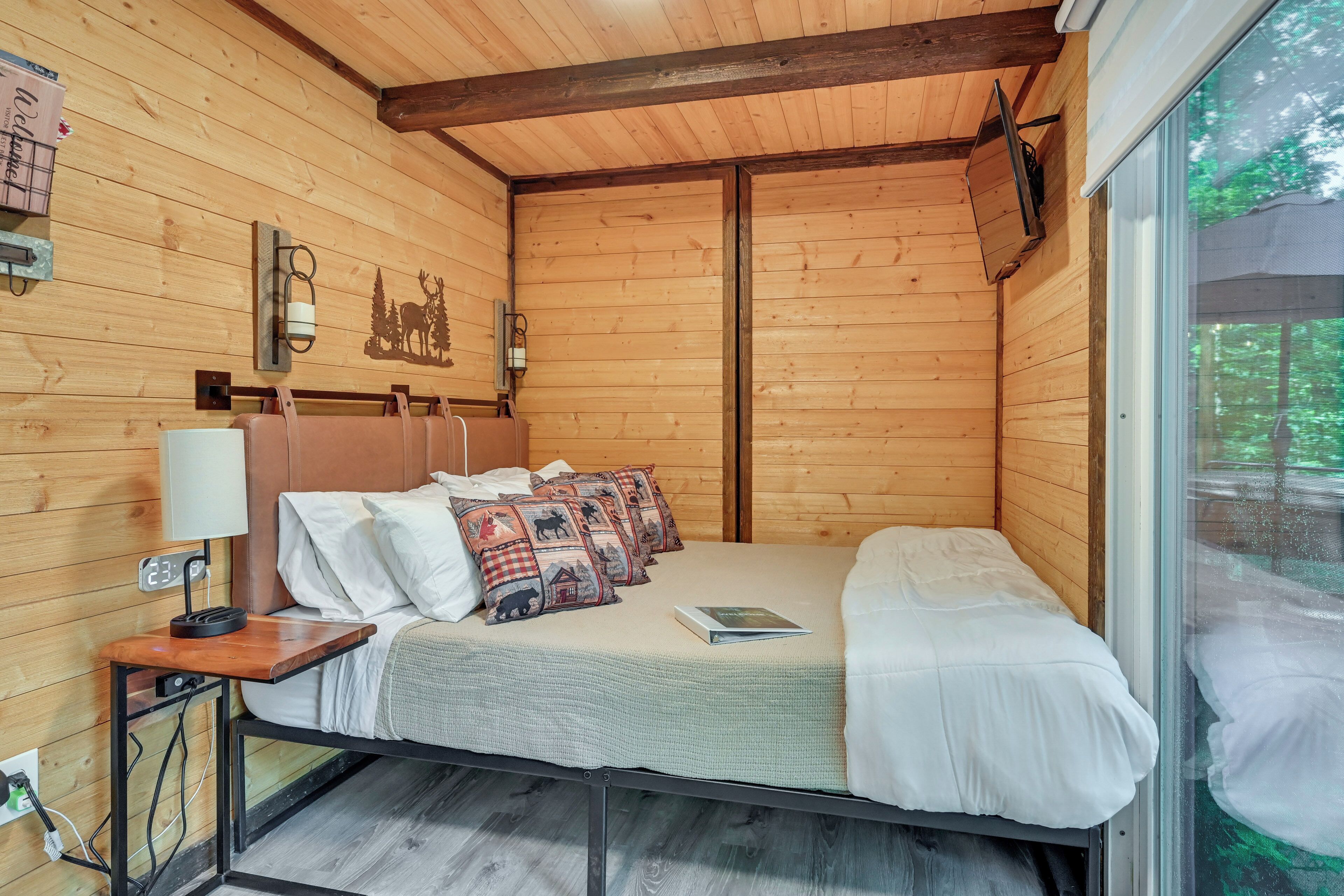 A wooden cabin room with a bed, pillows, a nightstand, a lamp, and a wall clock.