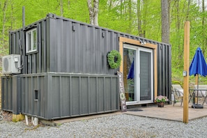 A compact, grey metal structure with a sliding glass door, a small window, and a welcome sign. - Tiny Home w/ Hot Tub & Mini Highland Cows! (Loudonville)