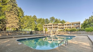 Une piscine entourée d’une terrasse, un immeuble à plusieurs étages doté de balcons, et une forêt dense en arrière-plan.