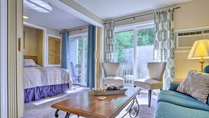 Une chambre à coucher avec un lit, un bureau et deux chaises. Il y a une fenêtre avec des rideaux, une lampe et une vue sur la verdure à l’extérieur.