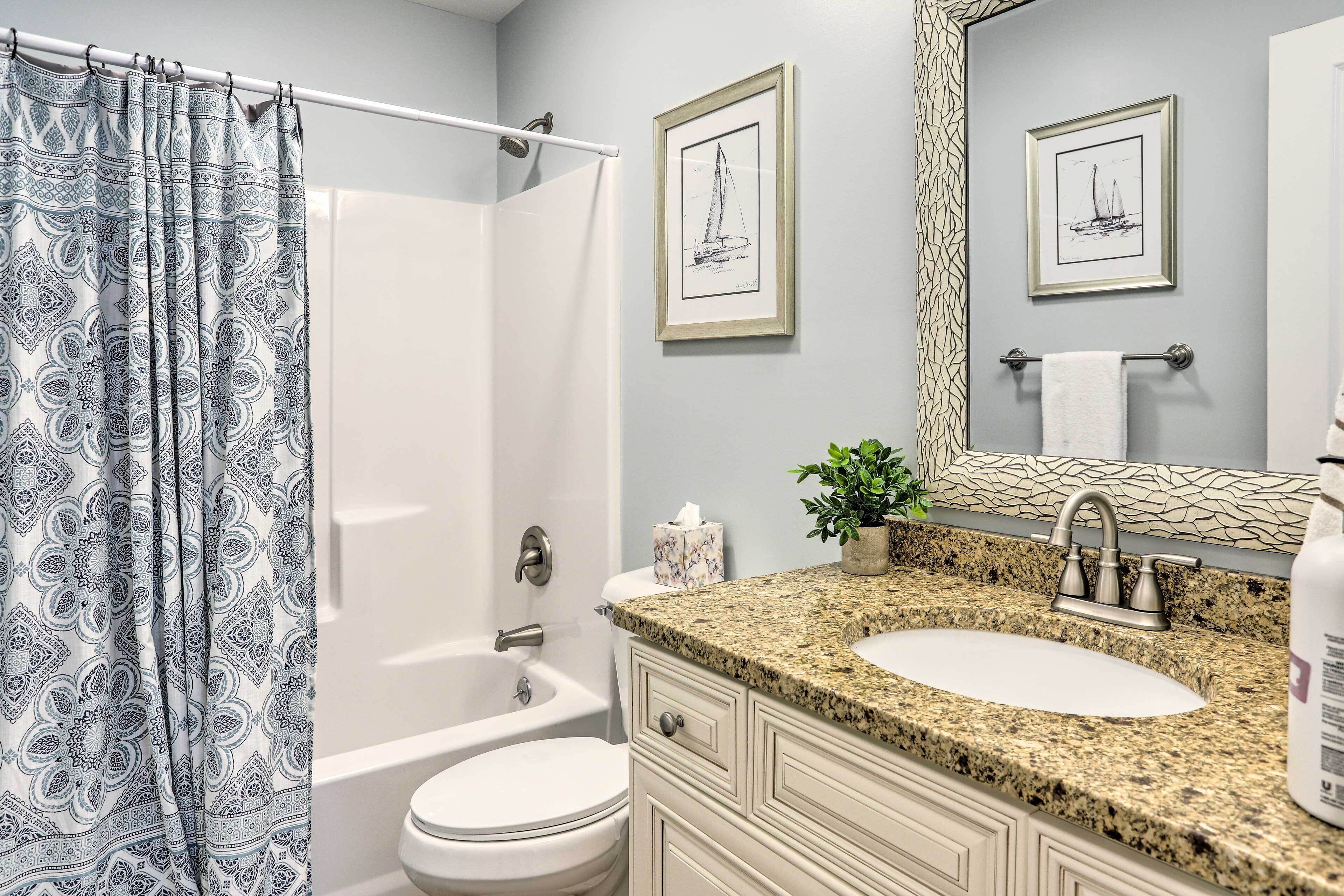 A bathroom with a shower curtain, a bathtub, a toilet, a sink with a granite countertop, and framed artwork on the wall.