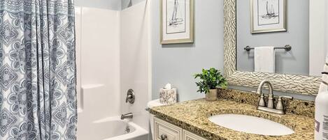 A bathroom with a shower curtain, a bathtub, a toilet, a sink with a granite countertop, and framed artwork on the wall.