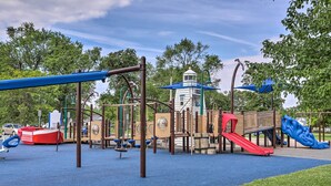 Ein Spielplatz mit verschiedenen Spielgeräten, darunter Rutschen und Schaukeln, umgeben von Bäumen und einem wolkenlosen Himmel.