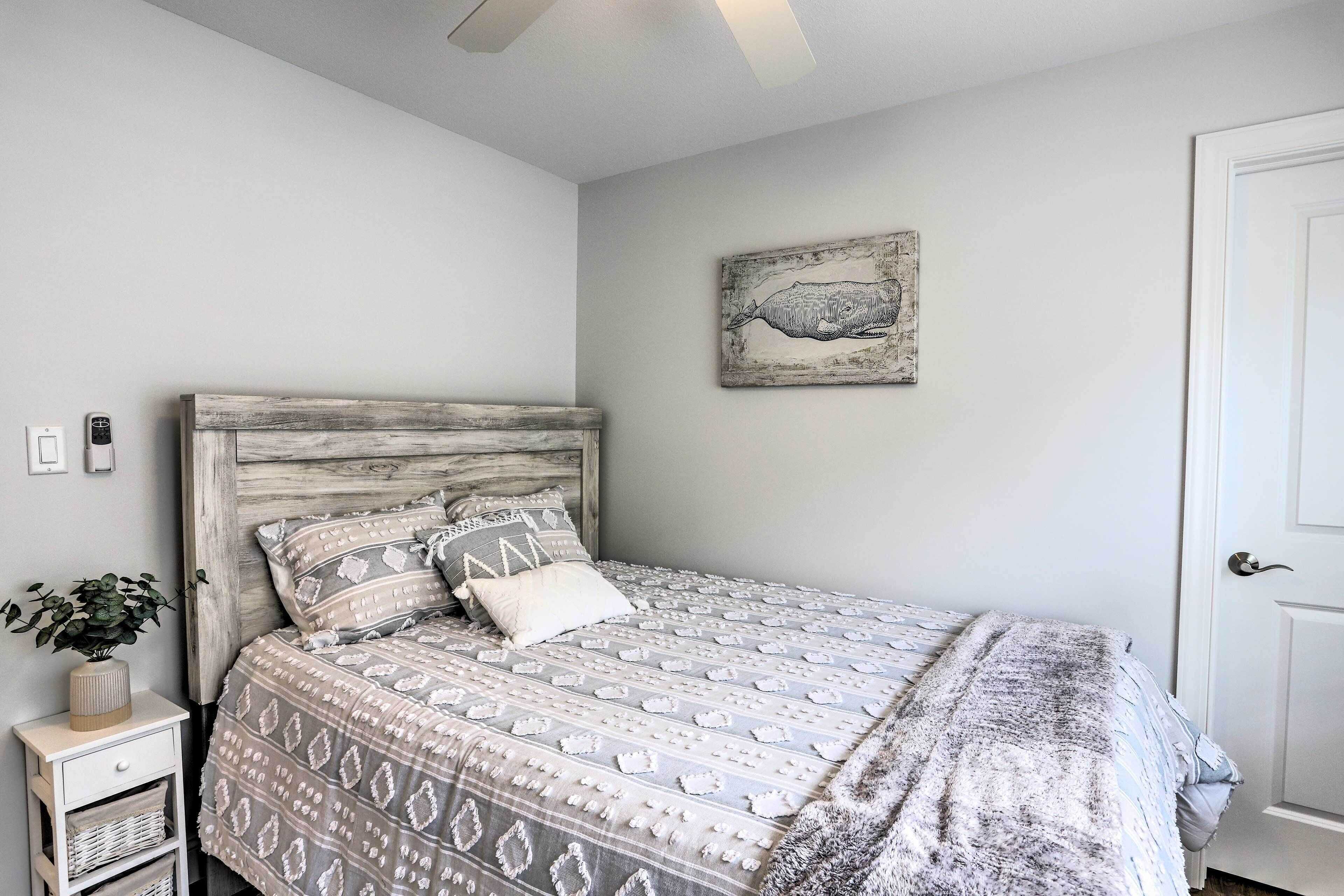 A bedroom with a wooden headboard, a patterned bedspread, a small bedside table with a plant, and a wall-mounted artwork.