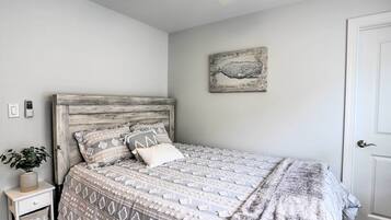 A bedroom with a wooden headboard, a patterned bedspread, a small bedside table with a plant, and a wall-mounted artwork.