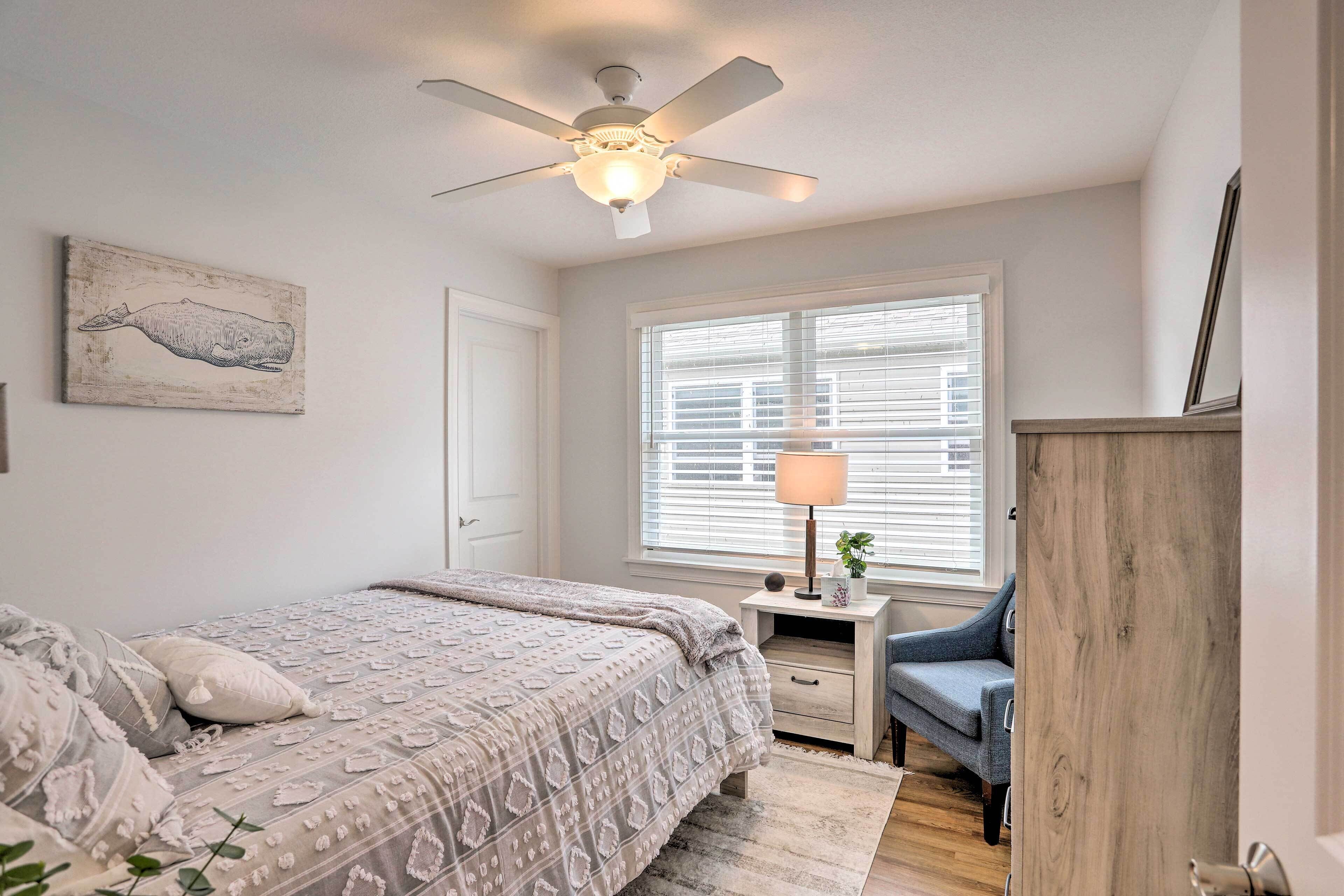 A bedroom with a bed, a ceiling fan, a window with blinds, a bedside table with a lamp, and a wall-mounted artwork.