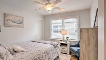 A bedroom with a bed, a ceiling fan, a window with blinds, a bedside table with a lamp, and a wall-mounted artwork.