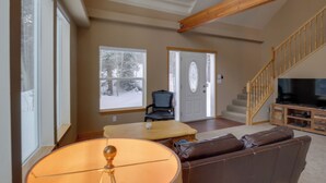 A spacious living room with a large window, a wooden staircase, a leather sofa, and a television.