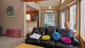 A cozy living room with a leather sofa, a wooden ceiling, and a large window with curtains.