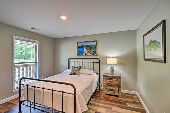 A bedroom with a metal bed frame, a bedside table with a lamp, a window with blinds, and a framed picture on the wall. - Bull Shoals Condo Retreat ~ 1 Mi to Marina! (Bull Shoals)