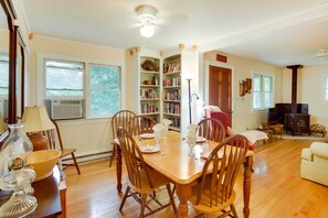 A dining area with a wooden table set for four, surrounded by chairs. There is a bookshelf filled with books and decorative items, a fireplace, and a cozy seating area with a sofa and armchairs. - Cozy 'nonny Cottage' Near Appalachian Trail! (Montebello)