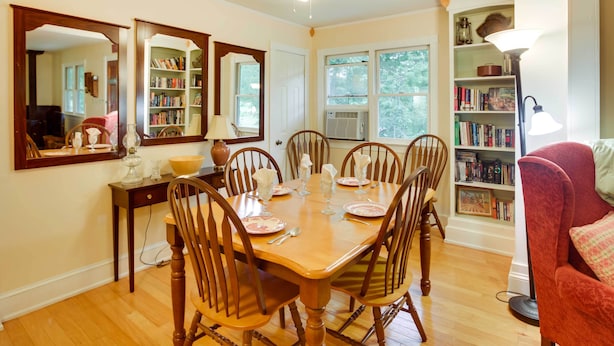 Une salle à manger avec une table en bois dressée pour quatre personnes, entourée de chaises. On y trouve une bibliothèque remplie de livres et d’objets décoratifs, une lampe et un miroir accroché au mur.