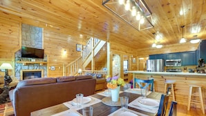 A wooden interior with a dining table set for four, a sofa, a stone fireplace, and a kitchen area.