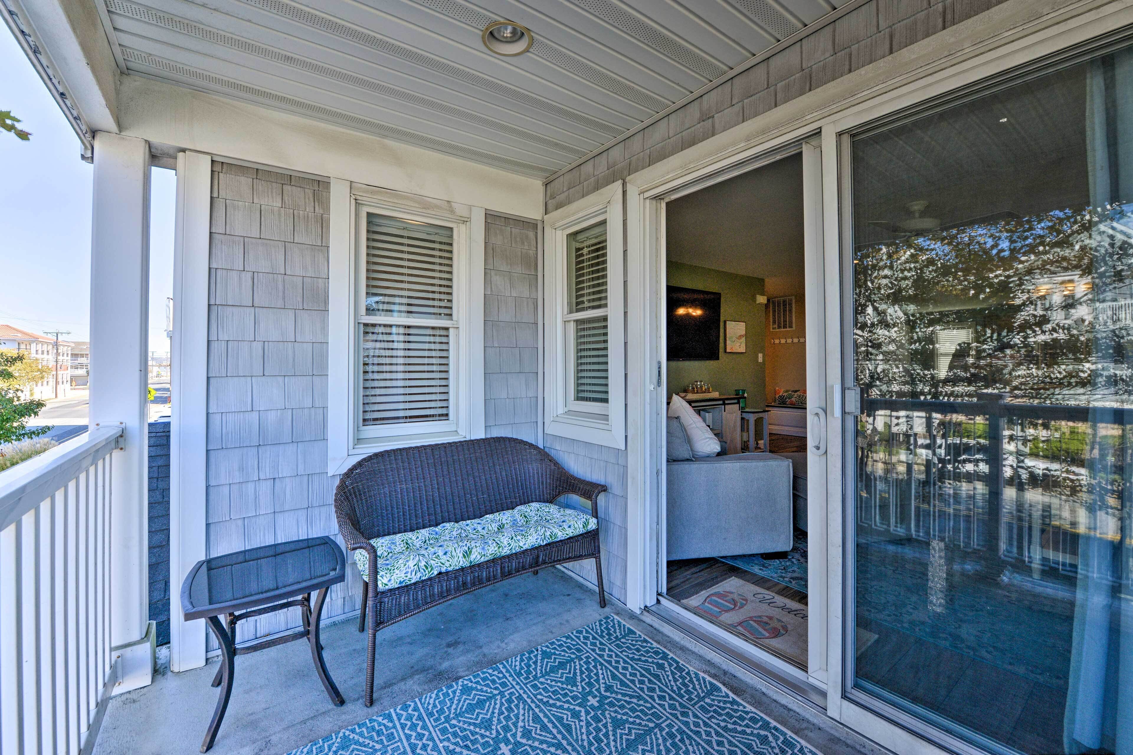 Un portico coperto con una panca in vimini e un tavolino, una porta scorrevole in vetro che dà su un patio, e un tappeto decorativo sul pavimento.