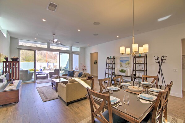 A spacious living area with a dining table set for six, a sofa, and a large window offering a view of the outdoors. - 068332: 'desert Pearl' 3 BR Oasis w/ Private Pool! (La Quinta)