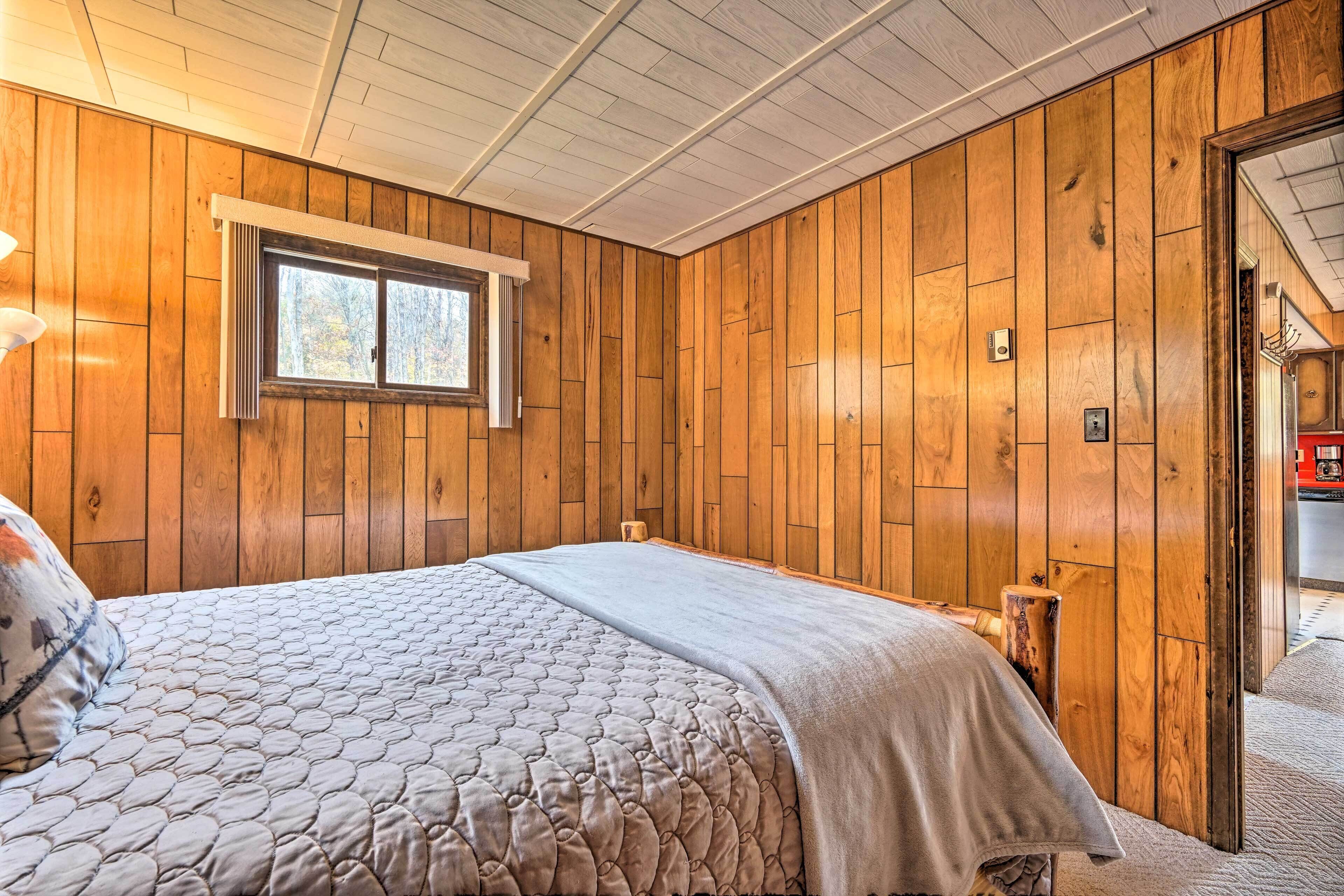 Une chambre avec des lambris en bois, un lit recouvert d’une courtepointe matelassée, une fenêtre et une porte donnant sur une autre pièce.
