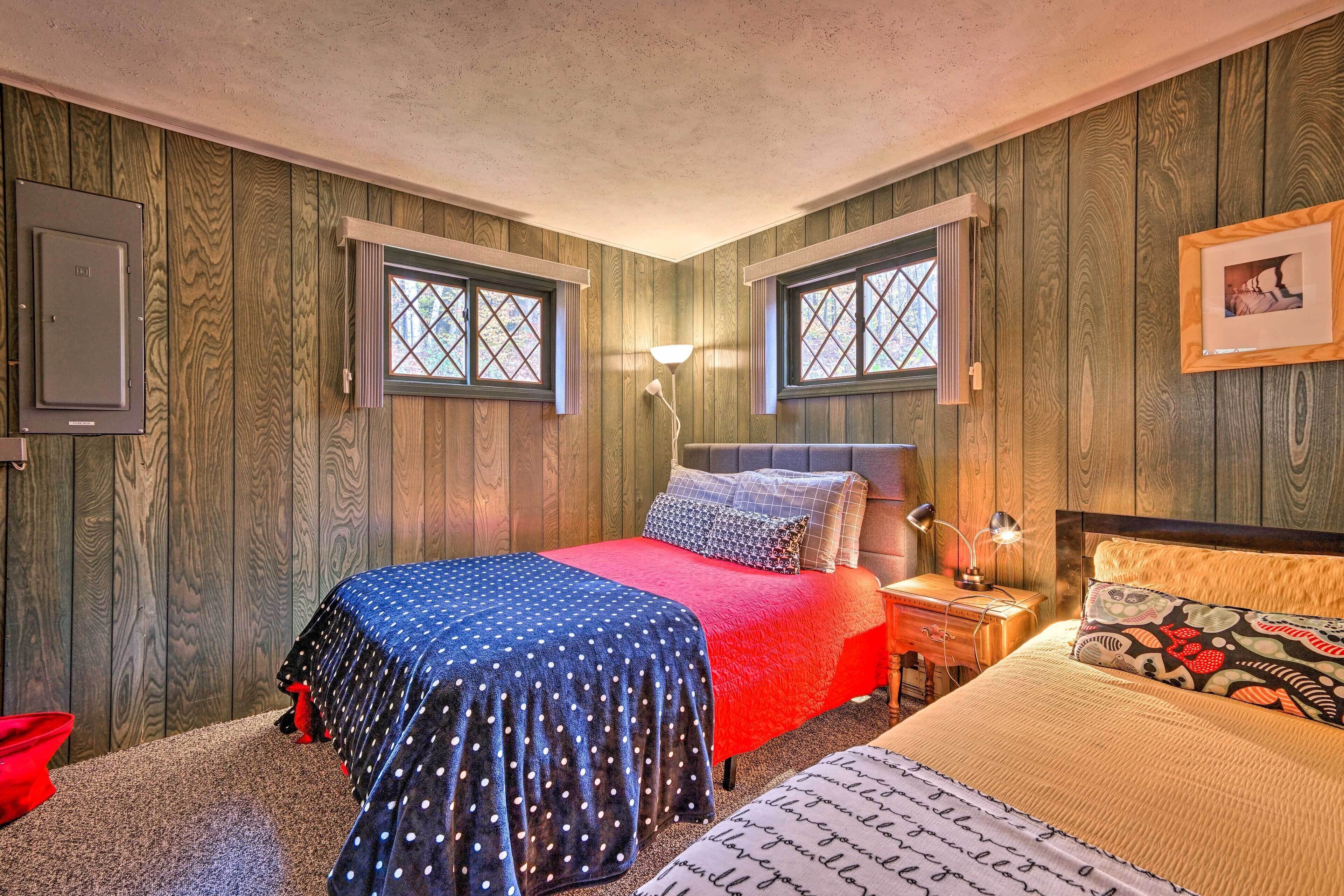 A bedroom with wooden walls, two windows, a bed with a polka dot and solid red bedspread, a nightstand with a lamp, and a framed picture on the wall.
