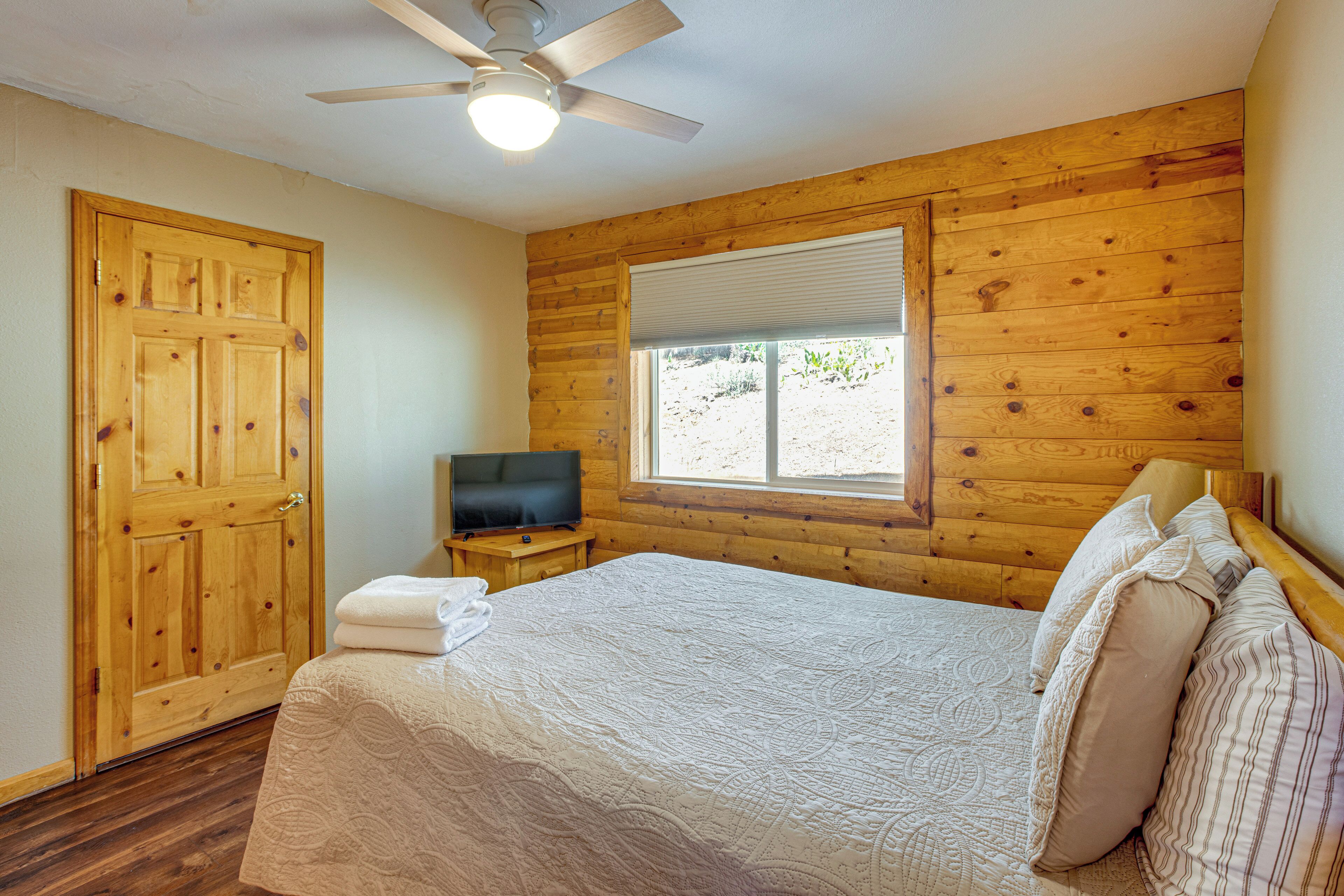 Un dormitorio con una pared de madera, una cama con almohadas, un televisor sobre una mesita de noche de madera y un ventilador de techo.