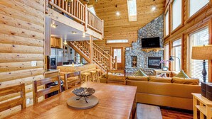 A log cabin interior with a dining area, a stone fireplace, a staircase, and a living room with a sofa.