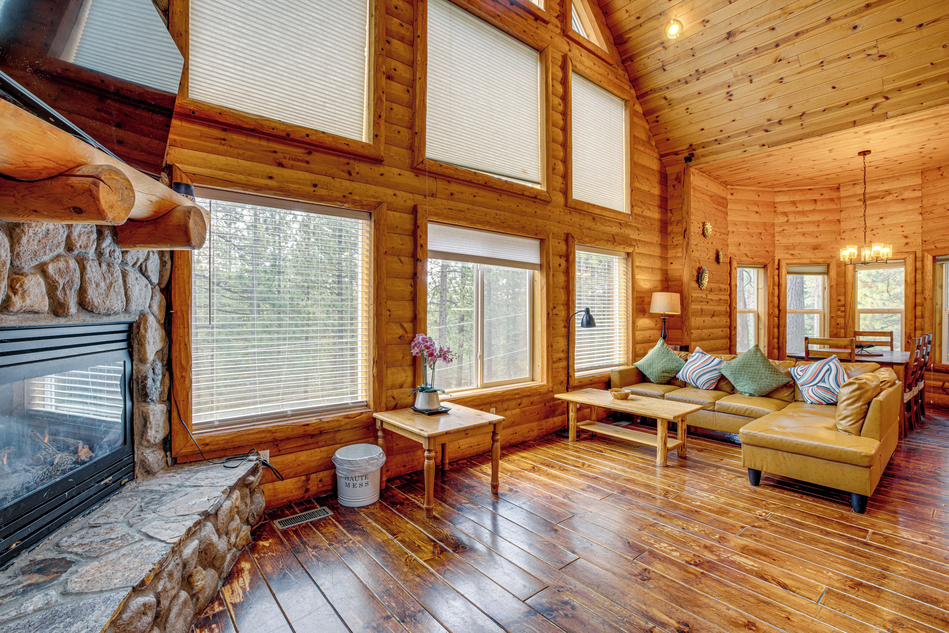 A log cabin interior with a stone fireplace, wooden floors, large windows, a sofa set, and a dining table.