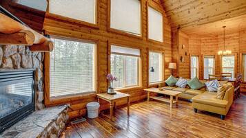 A log cabin interior with a stone fireplace, wooden floors, large windows, a sofa set, and a dining table.