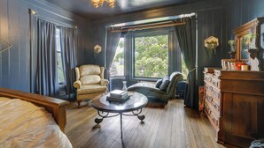 Een kamer met een bed, een ronde tafel met boeken, een stoel, een bank, een dressoir en een groot raam met gordijnen.