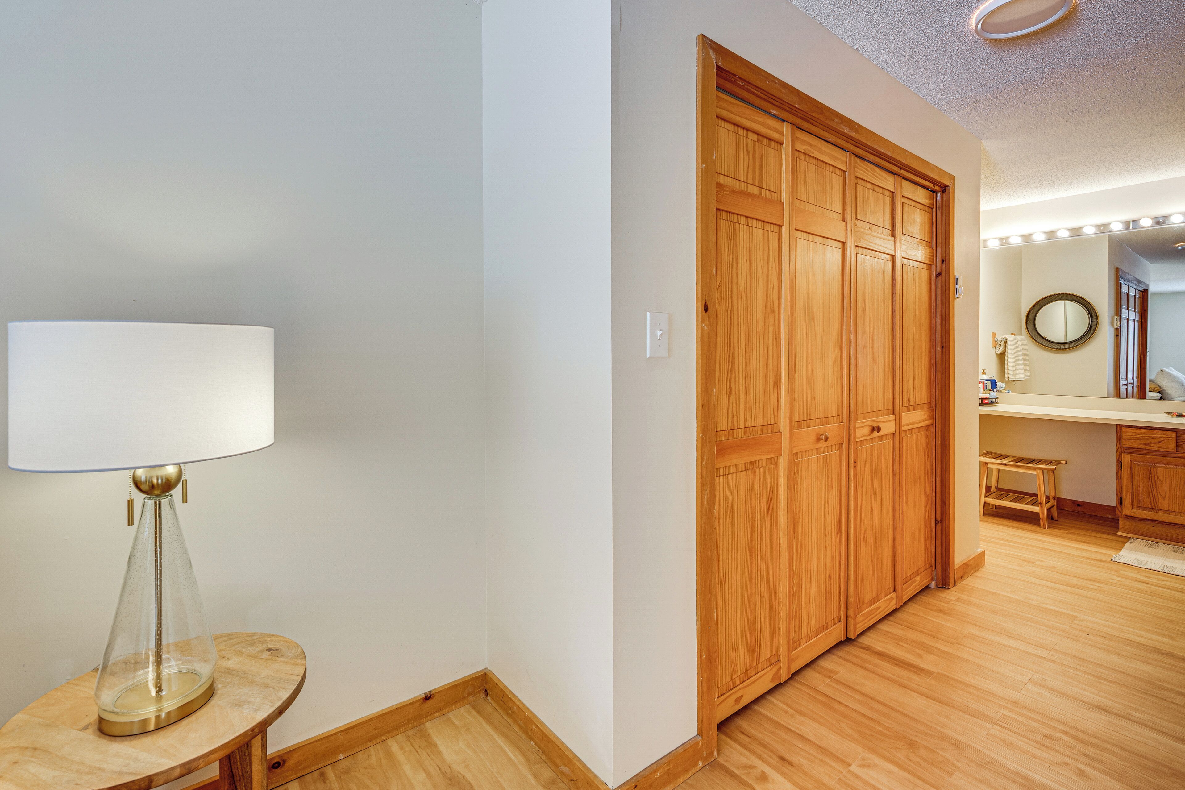 A hallway with a wooden wardrobe, a small table with a lamp, and a mirror with lights above the counter.