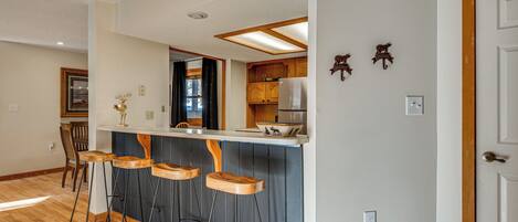 A kitchen with wooden bar stools, a refrigerator, and a window with curtains.
