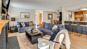 A modern living room with a sectional sofa, a coffee table, and a fireplace. There is a kitchen area with wooden cabinets and a bar stool.
