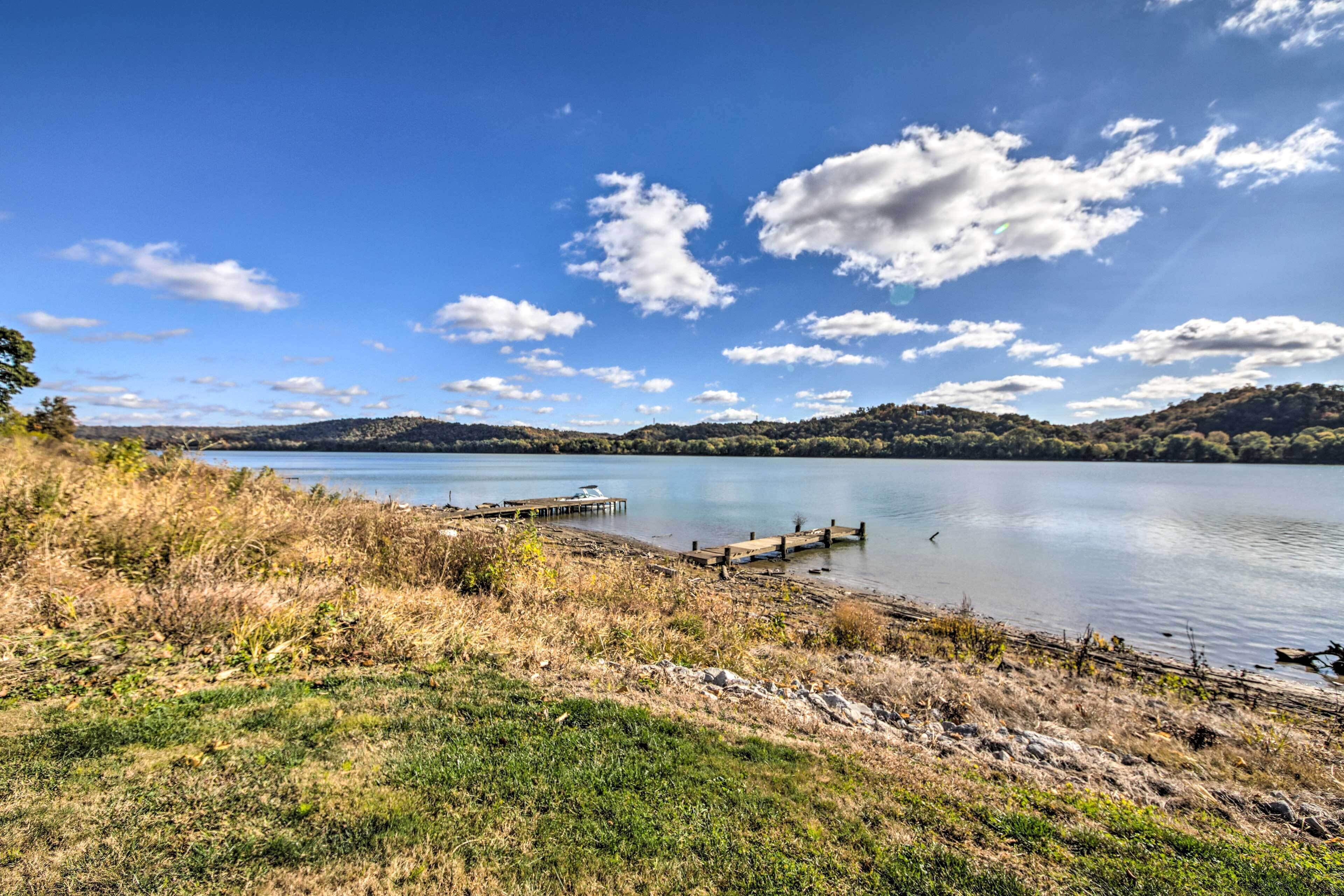 A lakeside view with a wooden dock extending into calm water, surrounded by grassy banks and hills in the background.