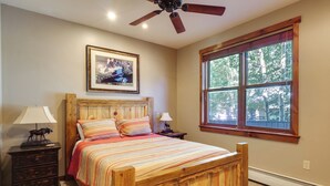 A bedroom with a wooden bed, a ceiling fan, a framed picture on the wall, and a large window with a view of trees. - Walk to Lifts: Breck Condo on Main St w/ Amenities (Breckenridge)