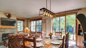 Un coin repas avec une table en bois dressée pour quatre personnes, entourée de chaises, d’une cheminée et d’un téléviseur fixé au mur.
