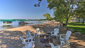 Ein Seeufer mit einer Holzterrasse, Adirondack-Stühlen, einer Feuerstelle und Blick auf Boote auf dem Wasser.