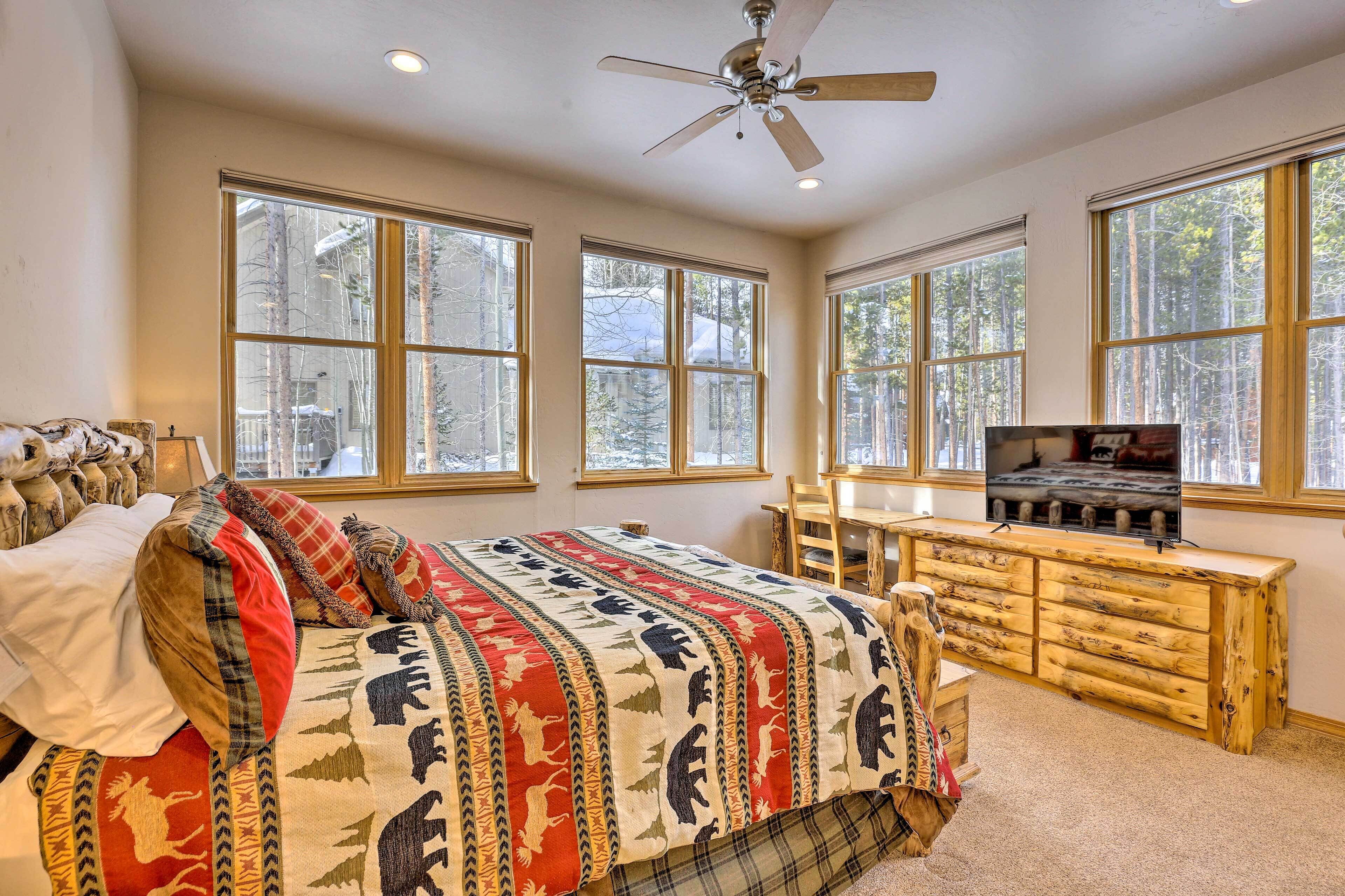 A bedroom with a large bed, a wooden dresser, and multiple windows offering a view of trees.