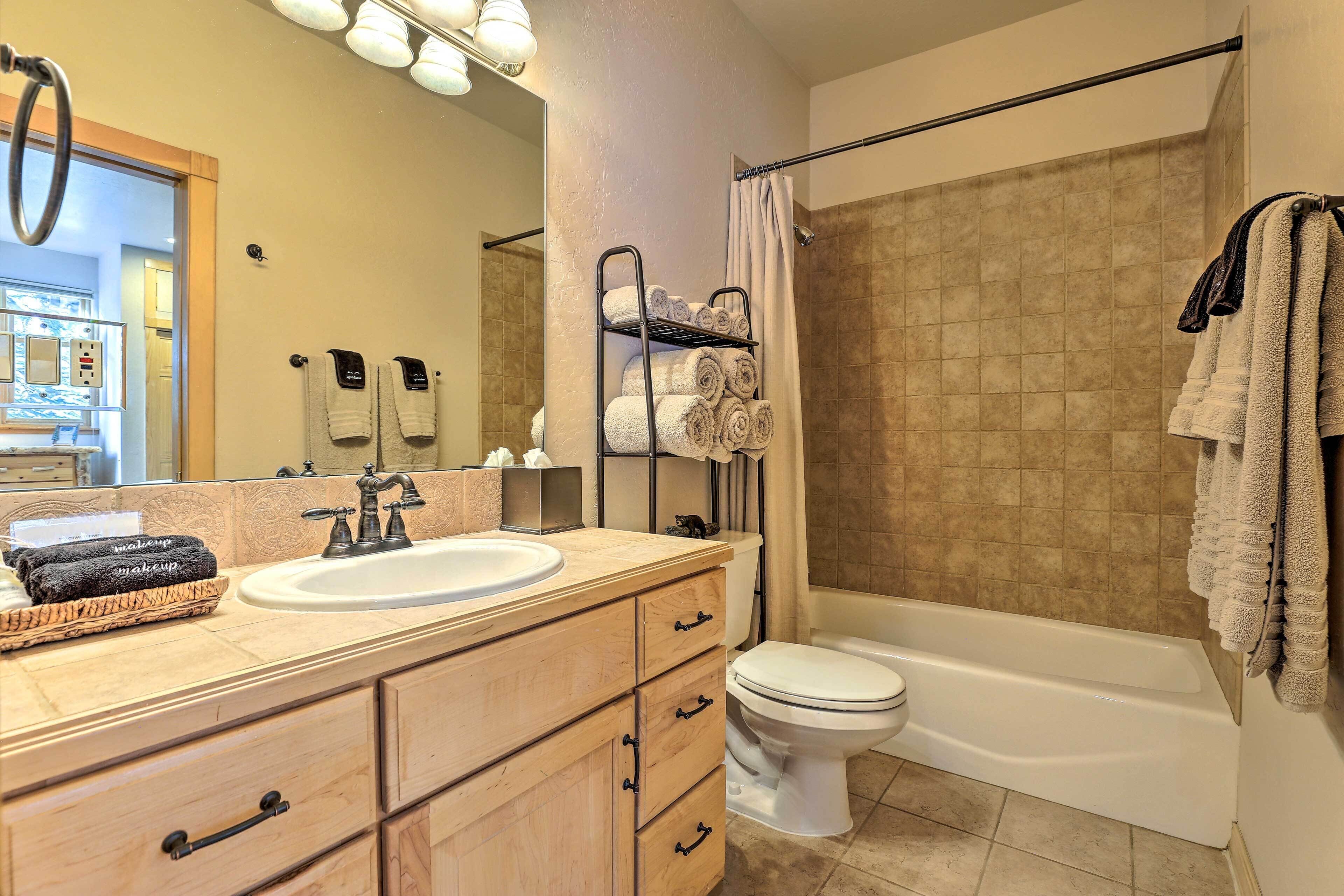 A bathroom with a sink, toilet, and bathtub. There are towels on a rack and a basket of makeup on the vanity.