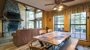 Una habitación amplia con chimenea de piedra, ventilador de techo de madera, ventanas grandes y una mesa de comedor con individuales.