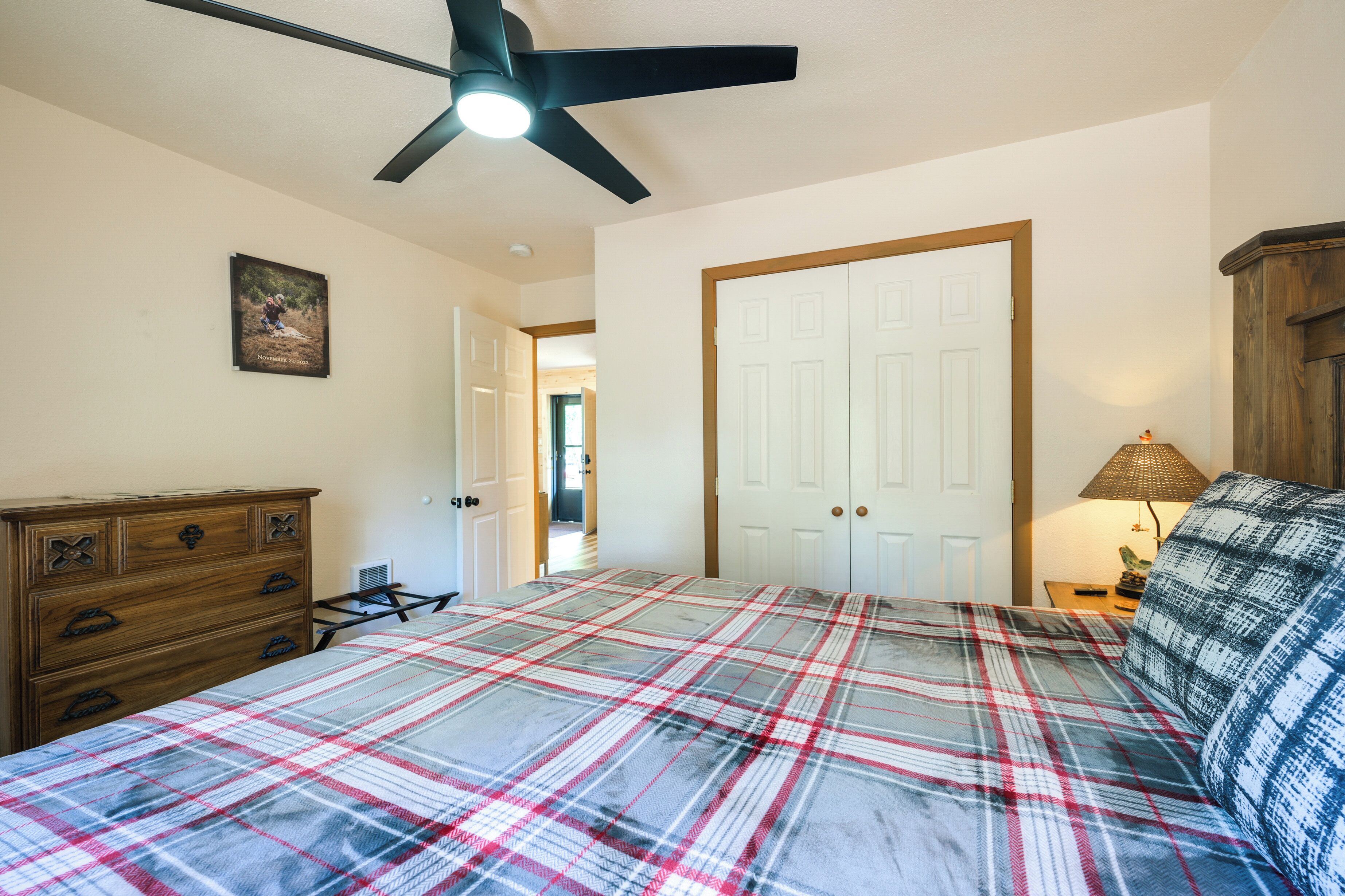 A bedroom with a plaid bedspread, a ceiling fan, a wooden dresser, and a door leading to another room.