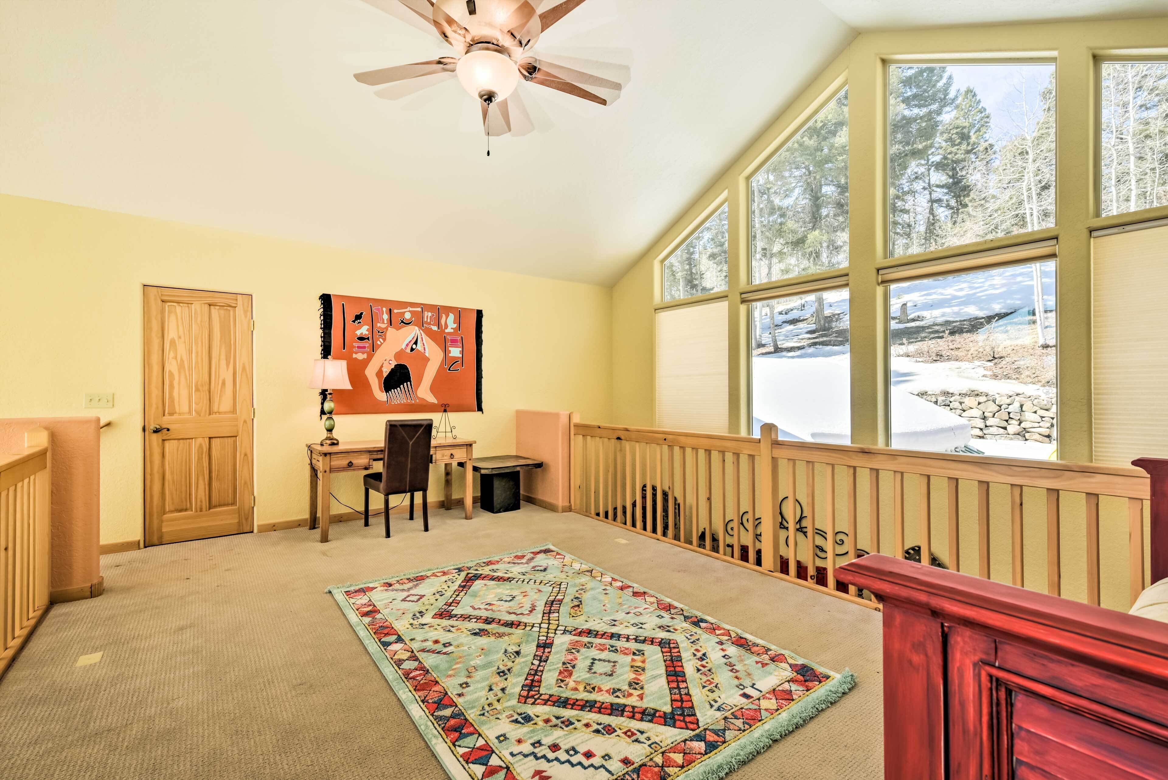 A room with a wooden desk, a chair, a rug, a ceiling fan, and large windows with a view of trees and snow.