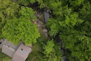 An aerial view of a house surrounded by dense green forest, with a visible rock formation and a small stream.