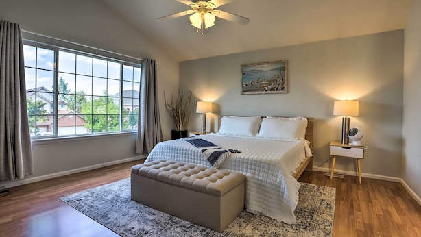 A bedroom with a large window, a ceiling fan, a bed with white bedding, a bedside table with a lamp, and a small nightstand with a fan. - Spacious Thornton Home: 15 Mi to Downtown Denver! (Thornton)