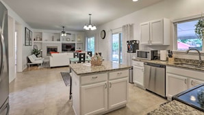 A modern kitchen with white cabinetry, granite countertops, and stainless steel appliances. There is a dining area with a white table and chairs, and a living room with a white sofa and a ceiling fan. - Spacious Thornton Home: 15 Mi to Downtown Denver! (Thornton)