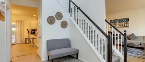 A staircase with white railings and a black handrail, leading to an upper floor. A gray bench is placed near the staircase, and a patterned rug is on the floor.