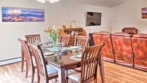 A dining area with a wooden table set for six, surrounded by chairs, a vase with flowers, and a mounted painting of mountains.