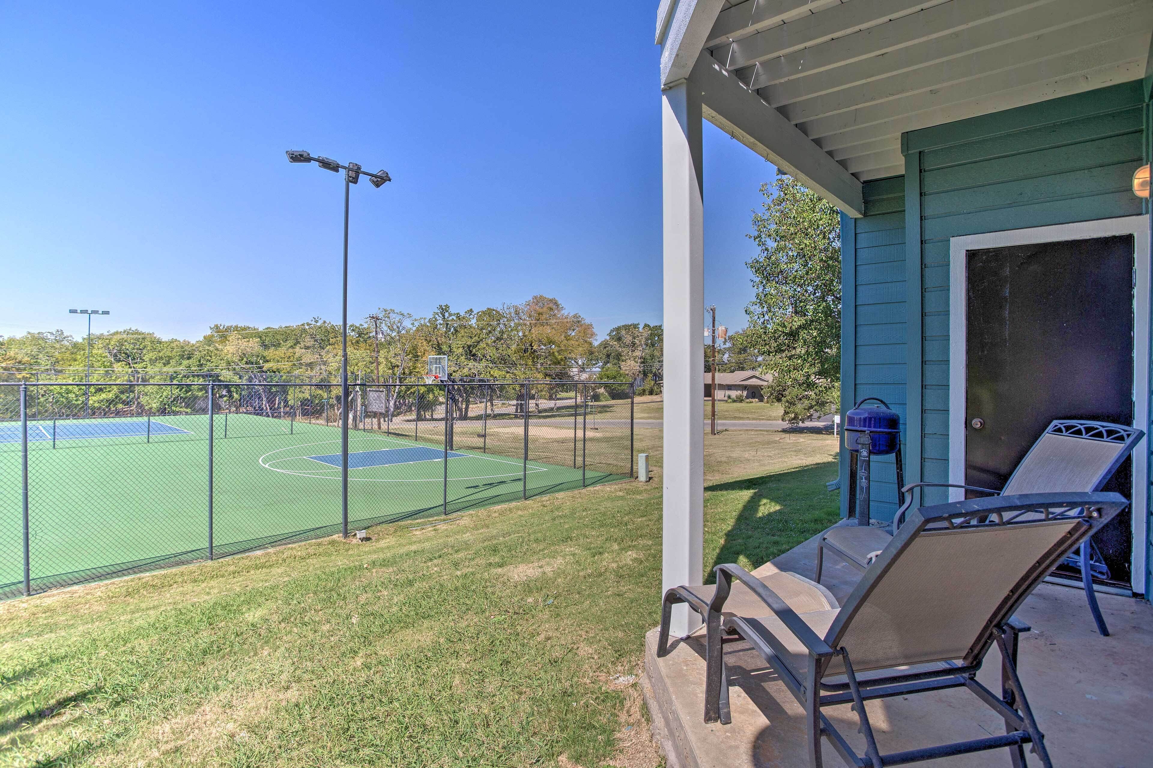 A tennis court with a net, surrounded by a fence, and a covered patio area with two chairs and a table.