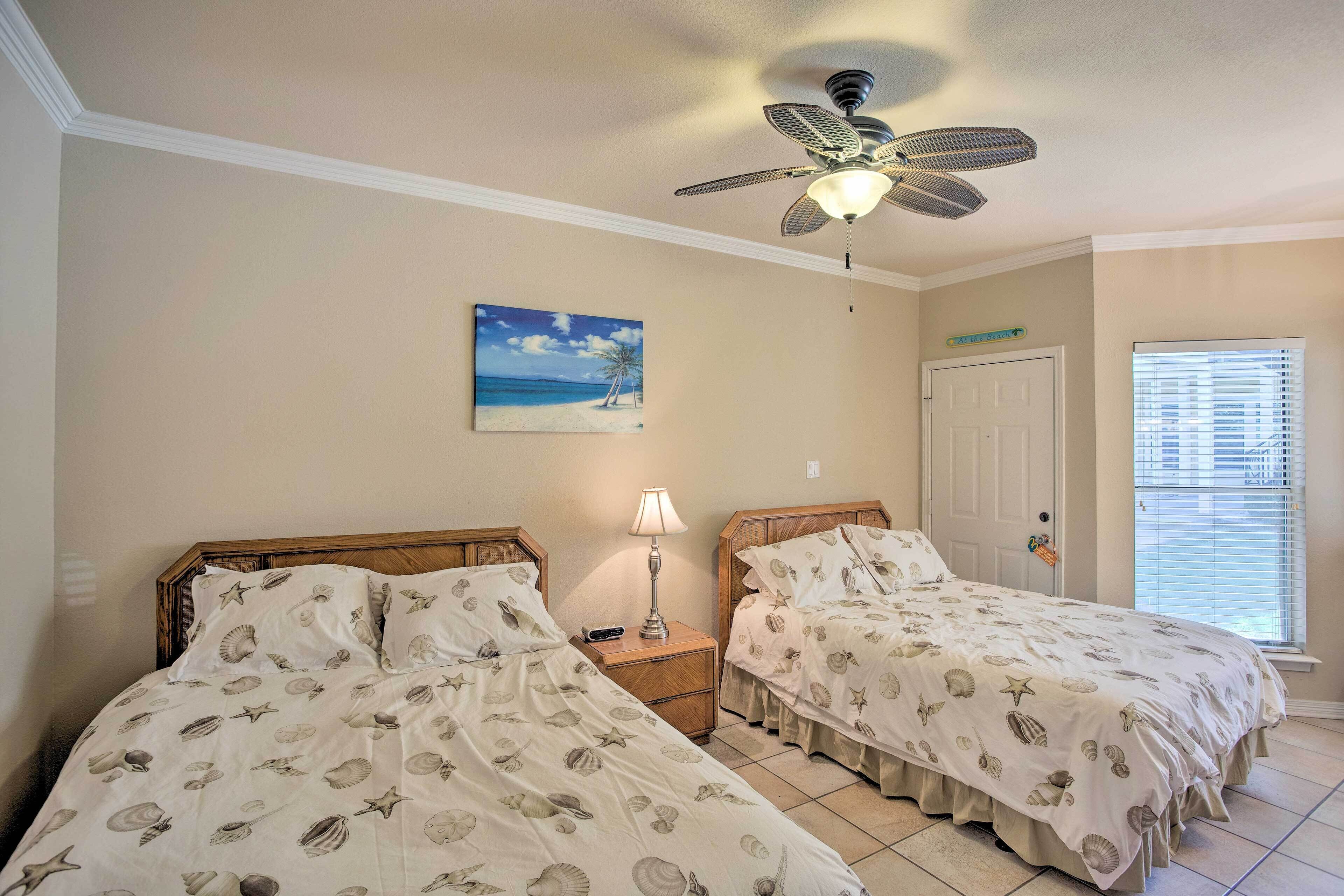 Un dormitorio con dos camas, un ventilador de techo, un cuadro de una playa y una puerta.