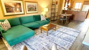 A living room with a green sofa, a coffee table, and a patterned rug. There is a dining area with a table and chairs. - 3 Bedroom Townhome in Osoyoos with lake views! XI (Osoyoos)