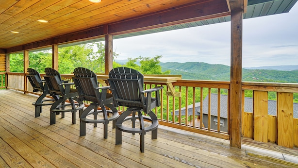 A wooden deck with four Adirondack chairs and a wooden table, overlooking a mountainous landscape.