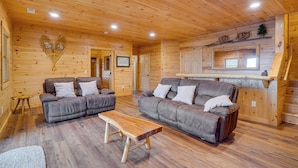 A wooden cabin interior with two sofas, a wooden coffee table, and a kitchen area with a window.